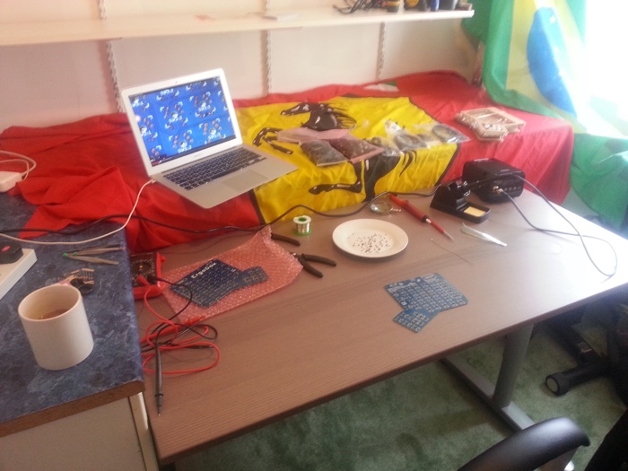 
   Photo of my desk during the assembly process.
   A Macbook with a fullscreen video showing a closeup of the circuit board is on top of a large red Ferrari flag alongside many bags of parts.
   On the desk itself are a soldering iron, several tools, a plate full of components, and the keyboard's circuit boards.