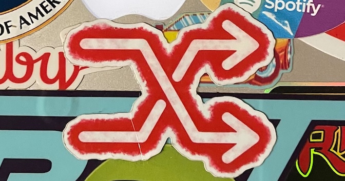 A shuffle symbol with a white foreground and a red spraypaint outline is seen affixed as a sticker to a laptop next to other stickers.
