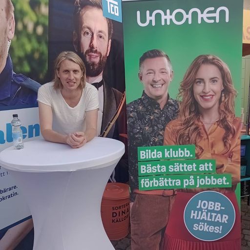 Person stood smiling at a booth at a conference. Next to them is a banner for the Swedish trade union “Unionen” with a slogan encouraging people to organise unions at their own workplaces.
