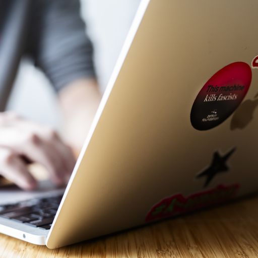 Laptop on table in the foreground, person in a grey hoodie typing in the background. A sticker reading "This machine kills fascists" is visible on the laptop.
